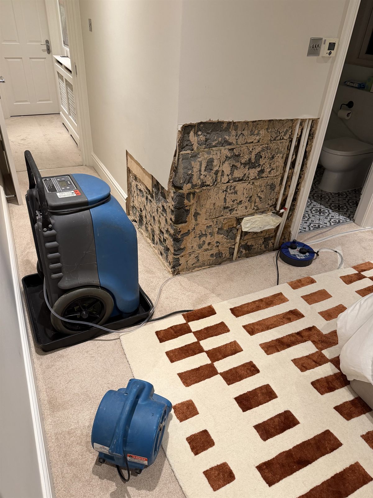 Bedroom wall stripped back after an en-suite leak, with a dehumidifier and air mover drying it